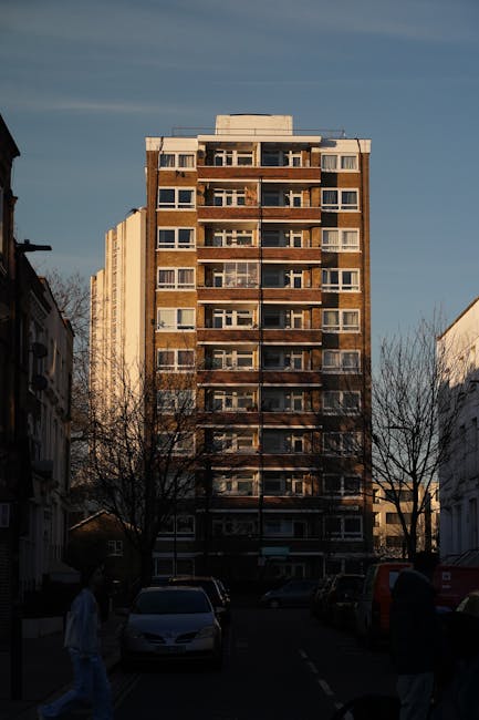 A multi-storey residential building with a brown brick façade and numerous balconies, situated at the end of a street with parked cars along the pavement. The sky above is partly cloudy, illuminated b