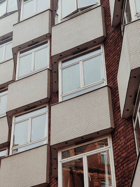 A multi-storey residential building with a brown brick façade and numerous balconies, situated at the end of a street with parked cars along the pavement. The sky above is partly cloudy, illuminated b