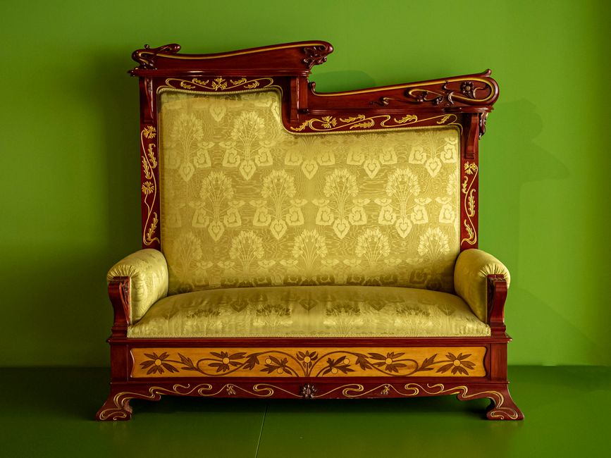 In a well-lit room with white walls and wooden ceiling beams, two movers from [COMPANY_NAME] are handling a home relocation. One individual, with curly hair and wearing a navy shirt, is positioned in 